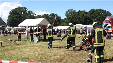 Feuerwehrleute in Einsatzkleidung führen bei einem Leistungsvergleich auf einer Wiese Übungsaufgaben mit Schläuchen und Geräten durch, während im Hintergrund Zuschauer unter Pavillons stehen.