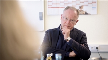 Ein Mann mit schwarzem Anzug und Brille schaut nachdenklich auf eine Person im Vordergrund.