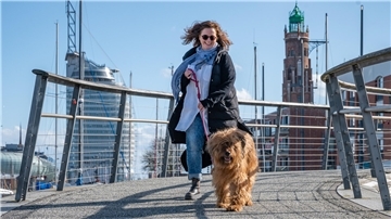 Eine Frau mit einem Briard-Hund lacht in die Kamera. Im Hintergrund sieht man das Atlantic Hotel Sail City in Bremerhaven.