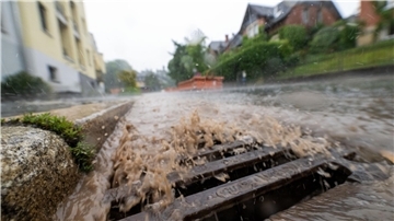 Starkregen überflutet eine städtische Straße; braunes Wasser schießt aus einem überlasteten Gully, im Hintergrund sind Wohnhäuser und Bäume erkennbar.