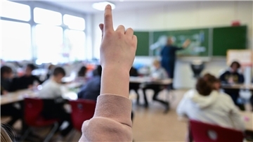 Unterrichtsszene: Ein Kind meldet sich, während der Lehrer vorn an der Tafel steht.