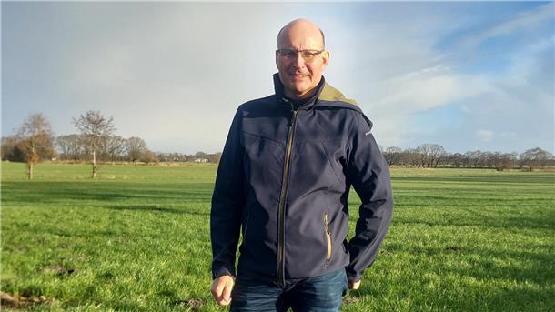 Ein Mann steht auf einer grünen Wiese, trägt eine dunkelblaue Jacke mit Reißverschluss und Jeans. Im Hintergrund sind Bäume und ein klarer Himmel mit vereinzelten Wolken zu sehen.