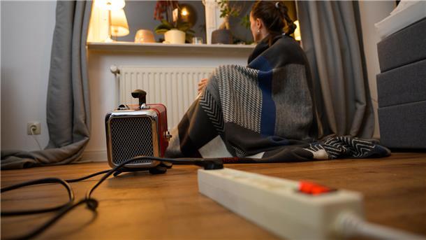 Ein Frau sitzt mit einer umgehangenen Decke hinter einem Elektro-Heizlüfter (gestellte Szene).
