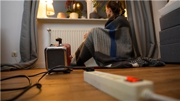 Ein Frau sitzt mit einer umgehangenen Decke hinter einem Elektro-Heizlüfter (gestellte Szene).