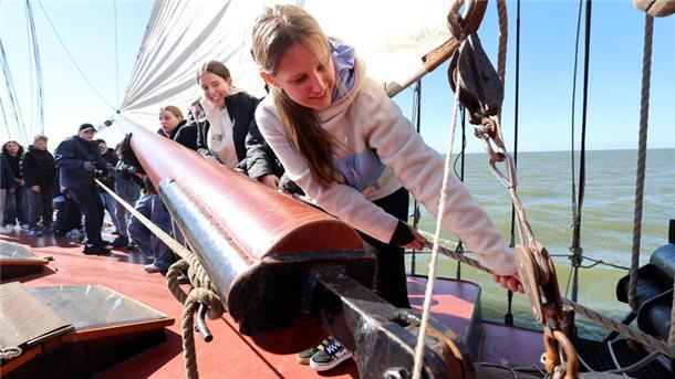 Mehrere Jugendliche stehen an Deck eines Segelschiffs und helfen aktiv beim Bedienen der Takelage; im Vordergrund zieht ein Mädchen konzentriert an einem Seil. Im Hintergrund ist das offene Meer zu sehen.