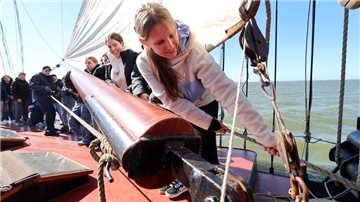 Mehrere Jugendliche stehen an Deck eines Segelschiffs und helfen aktiv beim Bedienen der Takelage; im Vordergrund zieht ein Mädchen konzentriert an einem Seil. Im Hintergrund ist das offene Meer zu sehen.