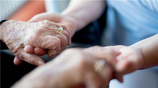 Eine Seniorin hält sich mit ihren Händen an den Händen einer Pflegekraft fest. 