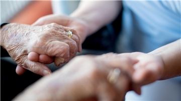 Eine Seniorin hält sich mit ihren Händen an den Händen einer Pflegekraft fest. 