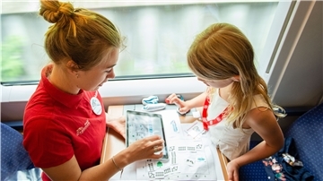 Eine junge Frau und ein kleines Mädchen sitzen sich in einem Zug gegenüber. Die Frau zeigt dem Mädchen etwas auf einem Tablet.
