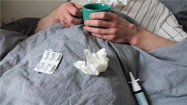 Ein Mann liegt mit Taschentüchern, Teetasse, Nasenspray und Tabletten im Bett.