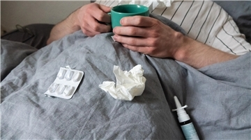 Ein Mann liegt mit Taschentüchern, Teetasse, Nasenspray und Tabletten im Bett.
