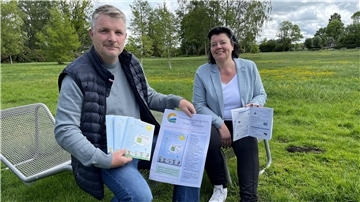 Zwei Personen sitzen auf einer Parkbank und präsentieren das Sommerferienprogramm der Stadt Geestland. Sie halten Broschüren und ein Plakat mit Programminhalten in den Händen. Im Hintergrund eine grüne Wiese mit Bäumen.