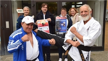 Vier Männer und zwei Frauen werben mit Plakaten und kleiner Showeinlage vor dem Eingang einer Sporthalle für eine Benefizveranstaltung.