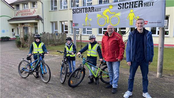 Drei Kinder mit Fahrrädern und reflektierenden Westen stehen vor einer Schule mit der Aufschrift 'Grundschule Neuenwalde'. Zwei Erwachsene, ein Mann in roter Jacke und ein Mann in blauer Jacke, stehen neben ihnen. Im Hintergrund hängt ein Banner mit der Aufschrift 'Sichtbarkeit = Sicherheit' und Grafiken von Personen und Fahrrädern. 