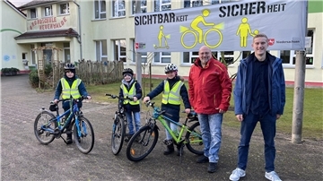 Drei Kinder mit Fahrrädern und reflektierenden Westen stehen vor einer Schule mit der Aufschrift 'Grundschule Neuenwalde'. Zwei Erwachsene, ein Mann in roter Jacke und ein Mann in blauer Jacke, stehen neben ihnen. Im Hintergrund hängt ein Banner mit der Aufschrift 'Sichtbarkeit = Sicherheit' und Grafiken von Personen und Fahrrädern. 