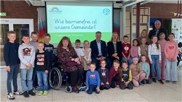 **ALT-Text:**
Gruppenfoto von Grundschulkindern, Lehrkräften, dem Bürgermeister und Mitgliedern des Inklusionsbeirats vor einer Präsentationsfolie mit der Aufschrift „Wie barrierefrei ist unsere Gemeinde? – Ergebnisse des Schülerrats“.