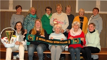 Gruppenfoto von 12 Frauen der Wiersdorfer Frauensportgruppe, die auf Bänken in einem Raum mit Holzverkleidung sitzen und stehen. Einige halten HSC-Fanutensilien wie einen Schal, einen Wimpel sowie einen Geschenkkorb.