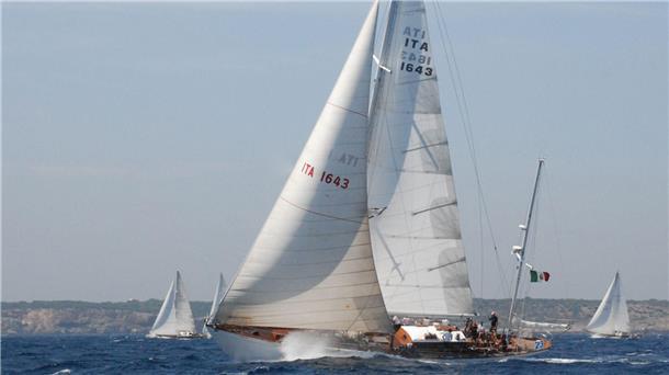 Die italienische Segelyacht „Corsaro II“ unter vollen weißen Segeln auf dem offenen Meer, mit sichtbarer italienischer Flagge am Heck. Weitere Segelschiffe sind im Hintergrund vor einer felsigen Küste zu sehen.
