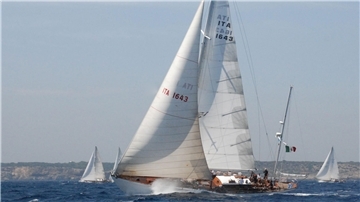 Die italienische Segelyacht „Corsaro II“ unter vollen weißen Segeln auf dem offenen Meer, mit sichtbarer italienischer Flagge am Heck. Weitere Segelschiffe sind im Hintergrund vor einer felsigen Küste zu sehen.
