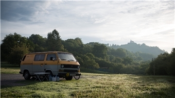 Ein VW Bulli steht auf einer Wiese vor einem Wald. Im Hintergrund ist eine Kirche auf einem Berg zu sehen.