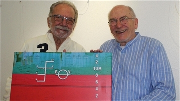 Zwei ältere Männer stehen lächelnd nebeneinander und halten gemeinsam ein farbenfrohes Foto oder Gemälde mit maritimem Motiv, das eine Schiffsbordwand mit Tiefgangmarkierungen und strukturierten Farbflächen zeigt.