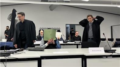 Ein Angeklagter sitzt in einem Gerichtssaal und hält sich eine grüne Mappe vor das Gesicht.