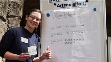 Eine Mitarbeiterin des Zoos am Meer steht lächelnd vor einer Tafel mit dem Titel „Unser Rekord“ und zeigt auf eine Liste gesammelter „Artendeckel“ der Jahre 2022 bis 2024.