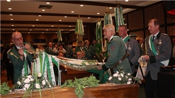 Mehrere Männer in grünen Uniformen stehen in einem Saal und präsentieren feierlich eine Fahne mit grünen Bändern. Einer von ihnen befestigt ein weiteres Band daran, während ein anderer die Fahne hält. Im Hintergrund sind weitere Uniformierte und Gäste in einem festlich dekorierten Raum zu sehen.
