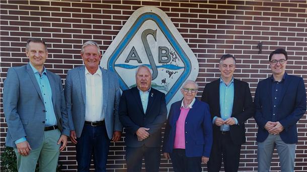 Engagement für Gewässer: Sportfischer Weser-Elbe feiern 50. Geburtstag Sechs Personen stehen in festlicher Kleidung vor einer Backsteinwand mit dem Logo einer Anglervereinigung mit den Buchstaben "ASB". Es handelt sich um Vertreter des Verbands und Ehrengäste bei einer Jubiläumsfeier.