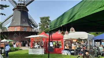 Marktszene vor der Seefelder Mühle: Mehrere bunte Verkaufsstände mit handgemachten Produkten auf einer Wiese, im Hintergrund die historische Windmühle bei blauem Himmel.