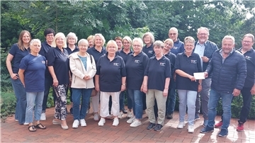 Gruppenfoto von rund 20 Mitgliedern des DRK-Ortsvereins Debstedt, die teils in dunklen DRK-Poloshirts gekleidet sind und im Freien auf einem gepflasterten Platz vor grünem Hintergrund stehen. Zwei Personen vorne rechts überreichen einen Umschlag.