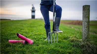 Eine Person trägt blaue Gummistiefel, daneben liegen pinke Stiefel im Gras. Im Hintergrund sieht man einen schwarz-weiß gestreiften Leuchtturm.
