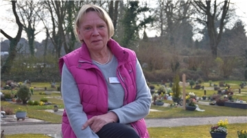 Karin Miosga sitzt auf einem der Bänke des Friedhofs in Atens. Im Hintergrund sind die Gräber zu sehen. 