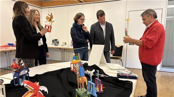 Papierkünstler Walter Ruffler zeigt mehreren interessierten Erwachsenen bunte mechanische Papierfiguren auf einer Tischpräsentation in einem Ausstellungsraum.