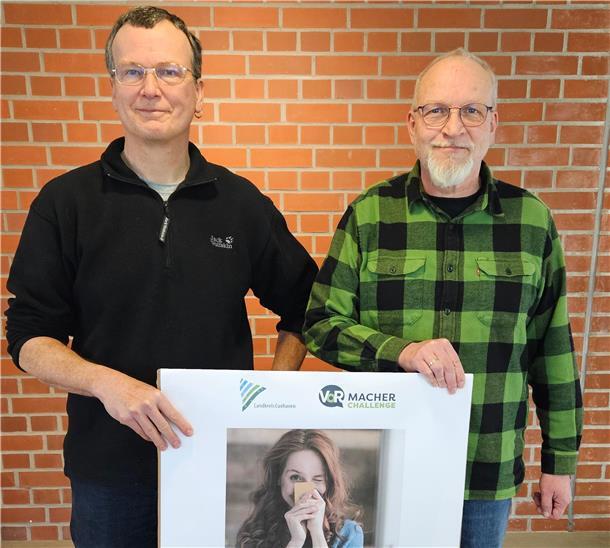 Zwei Männer mittleren Alters stehen vor einer roten Backsteinwand und halten gemeinsam ein großes Plakat. Der Mann links trägt eine schwarze Fleecejacke, der Mann rechts ein grün-schwarz kariertes Hemd. Das Plakat zeigt eine junge Frau mit langen Haaren, die eine gelbe Karte vor ihr Gesicht hält und lächelt. Oben auf dem Plakat sind die Logos des „Landkreis Cuxhaven“ und der „VoR Macher Challenge" zu sehen.