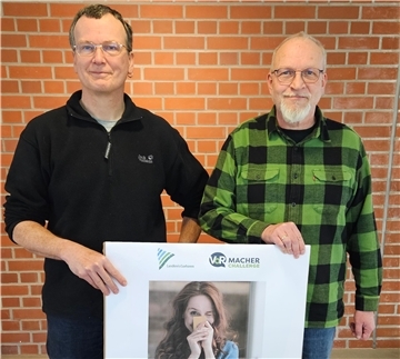 Zwei Männer mittleren Alters stehen vor einer roten Backsteinwand und halten gemeinsam ein großes Plakat. Der Mann links trägt eine schwarze Fleecejacke, der Mann rechts ein grün-schwarz kariertes Hemd. Das Plakat zeigt eine junge Frau mit langen Haaren, die eine gelbe Karte vor ihr Gesicht hält und lächelt. Oben auf dem Plakat sind die Logos des „Landkreis Cuxhaven“ und der „VoR Macher Challenge" zu sehen.