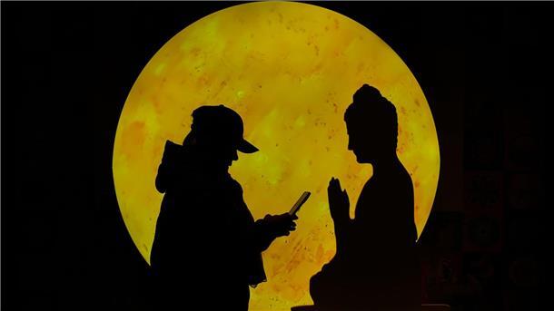 „Zwiegespräch“:  Eine Frau surft auf ihrem Smartphone vor einer Buddha-förmigen Pappfigur, die in einem Einkaufszentrum in Peking aufgestellt ist.